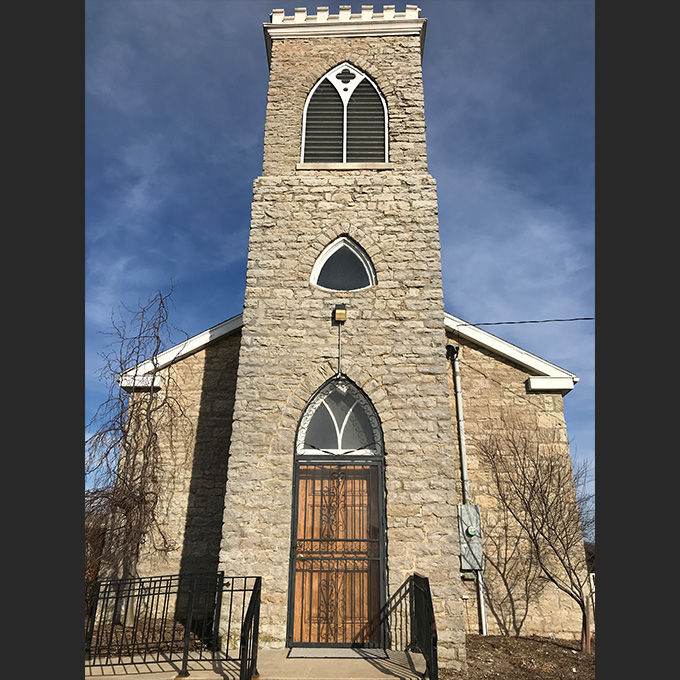 Front view of a stone church with a tall rectangular tower, arched windows, and a heavy wooden door behind an iron gate against a clear blue sky.